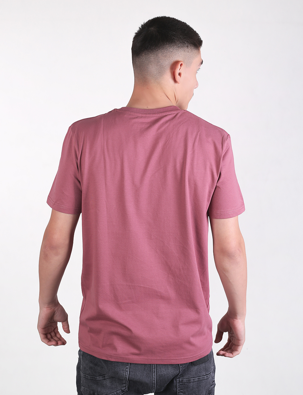 A young man with a short fade haircut is shown from behind against a plain off-white background. He is wearing a plain, mauve or dusty pink crew-neck t-shirt and dark grey or black denim jeans.