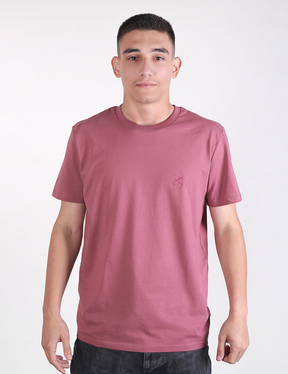 A young man with short dark hair standing against a plain white background, wearing a mauve crew-neck T-shirt and dark pants.