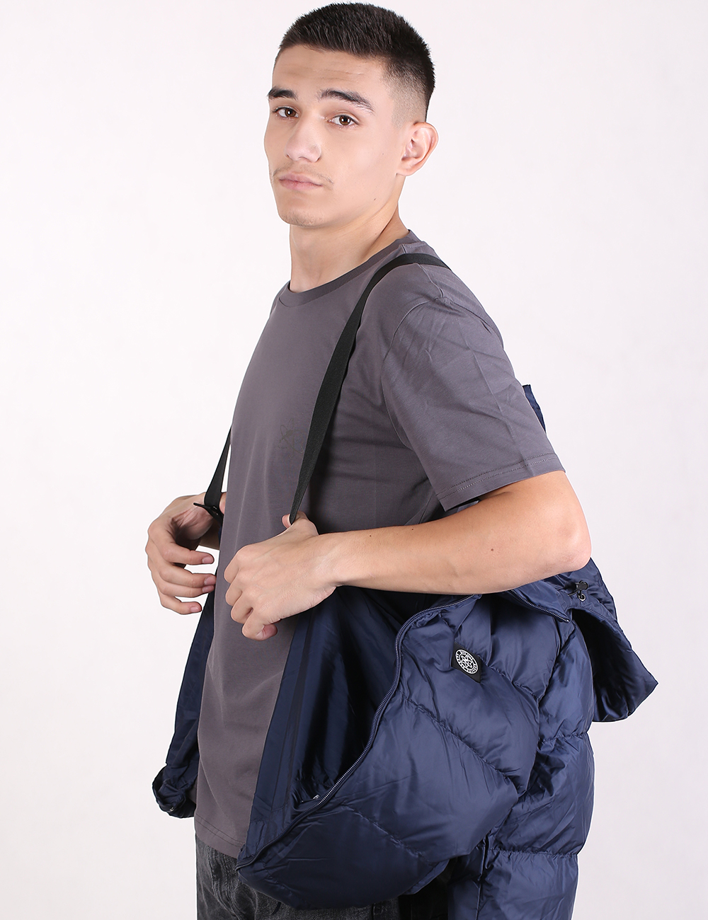 A man wearing a gray t-shirt and dark pants, carrying a navy blue puffer jacket over his shoulder with a matching shoulder bag strap visible.