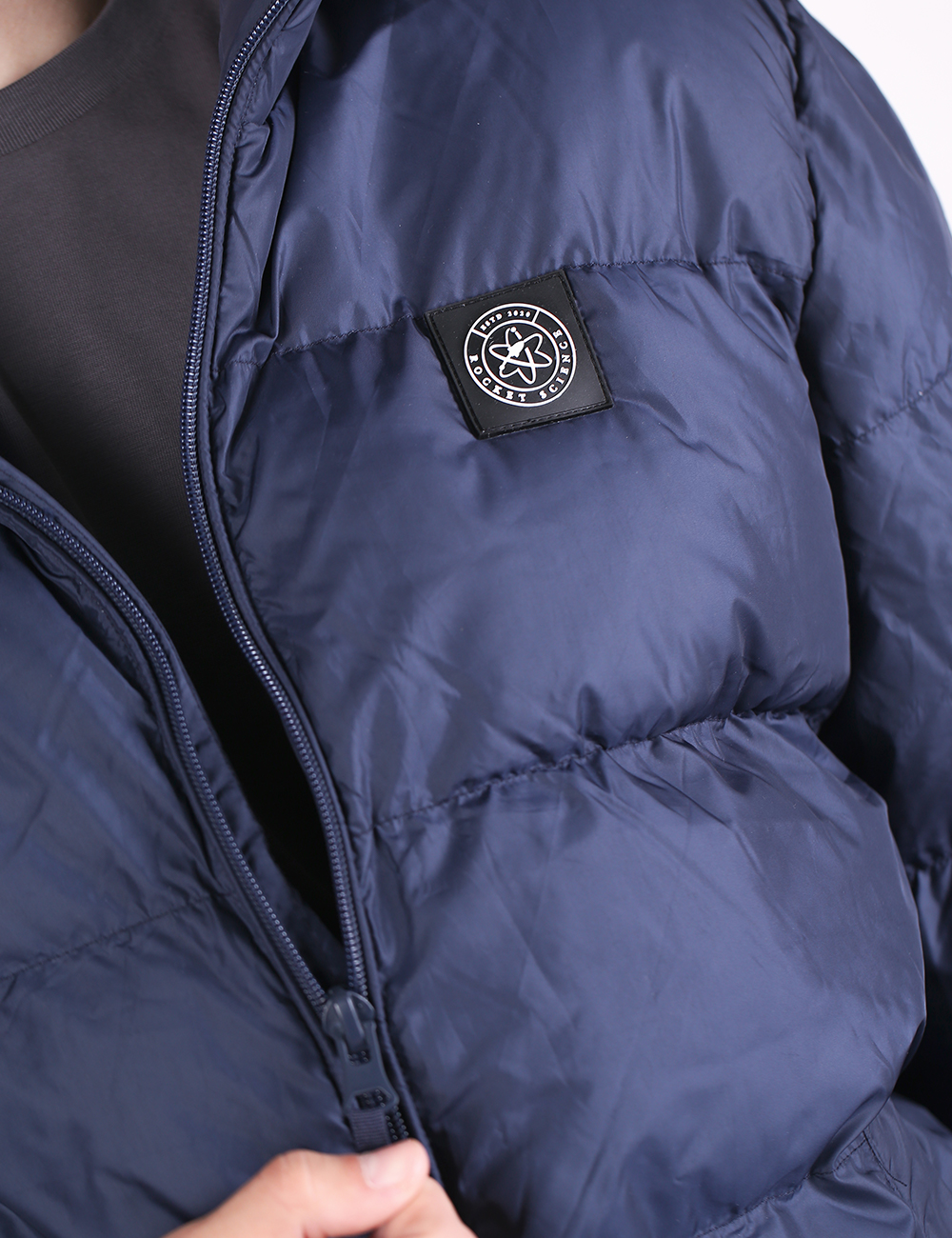 Close-up of a navy blue puffer jacket with horizontal quilting, a black zipper, and a black brand patch on the chest. A hand is visible at the bottom edge.