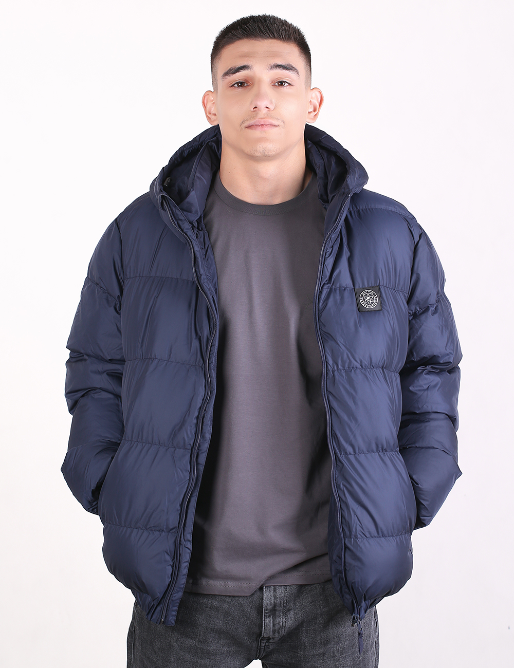 A man wearing a navy blue hooded puffer jacket over a dark gray t-shirt and dark jeans, standing against a plain white studio background.