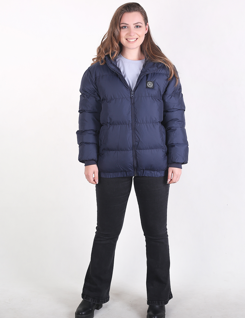 A woman modeling a navy blue puffer jacket with horizontal quilting, a front zipper, and a small circular chest logo, paired with black pants and a light gray top, against a plain white studio background.