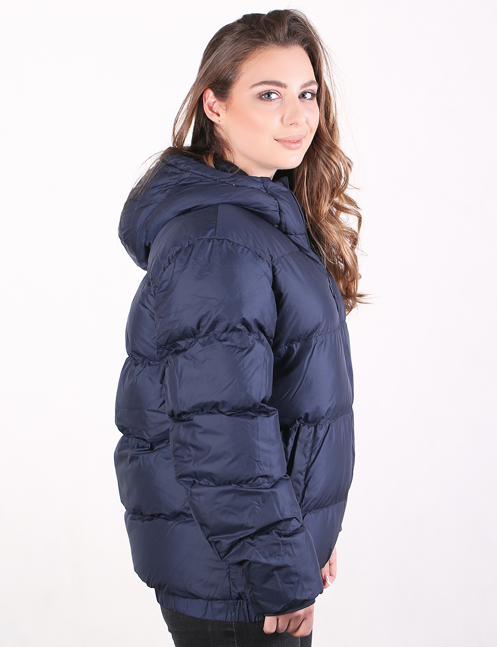 A woman wearing a navy blue hooded puffer jacket, shown from a side-back angle against a plain white studio background.