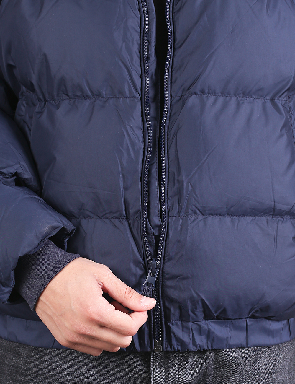 Close-up of a navy blue puffer jacket being zipped, with a person's hand holding the zipper pull and dark gray jeans visible below.