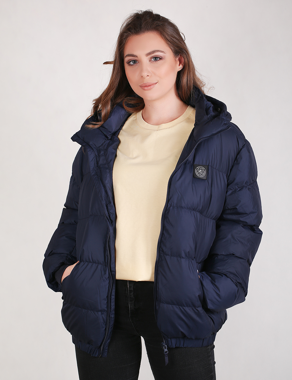 A woman wearing a navy blue puffer jacket over a cream-colored top and black pants, standing against a light gray studio background.