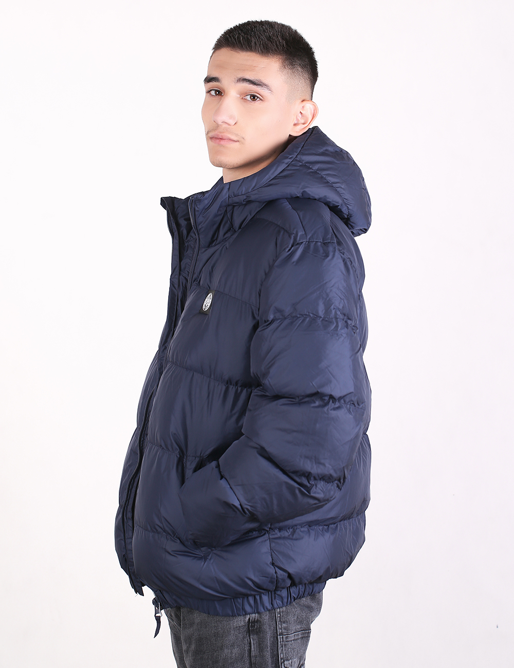 A man wearing a navy blue hooded puffer jacket, shown from a side profile against a plain white studio background.