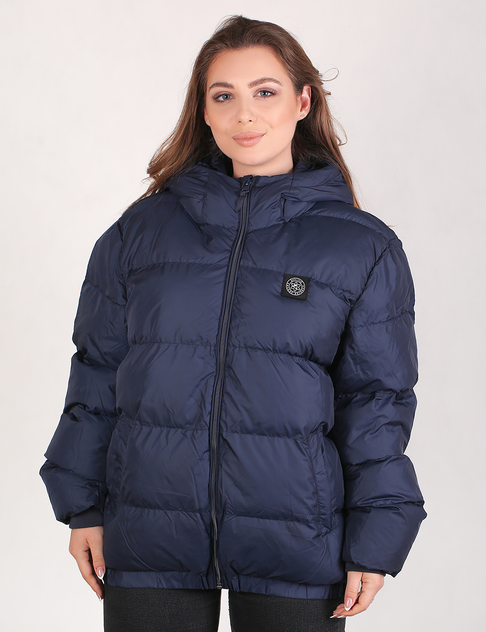 A woman wearing a navy blue puffer jacket with a black zipper and a circular black-and-white logo patch on the left chest, photographed against a light gray studio background.