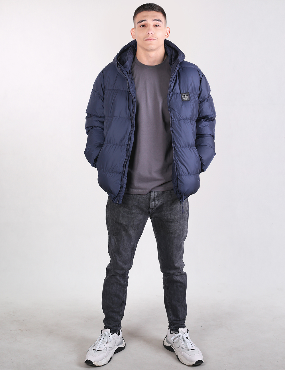 A man modeling a navy blue puffer jacket, worn over a dark gray t-shirt, dark gray jeans, and white sneakers, standing against a light gray studio background.