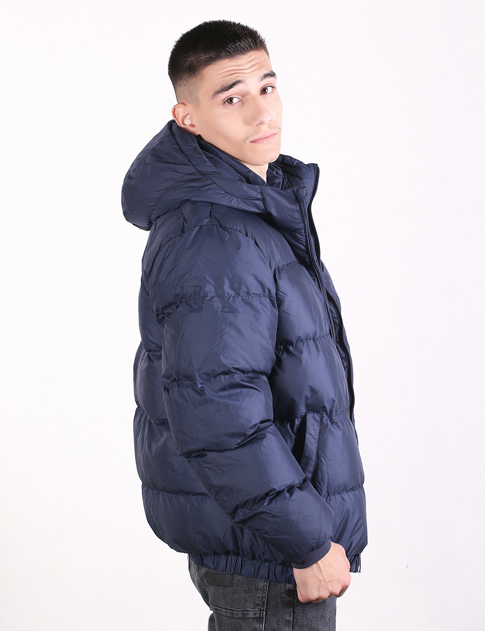 A man wearing a navy blue hooded puffer jacket, shown from a side profile against a plain white studio background.