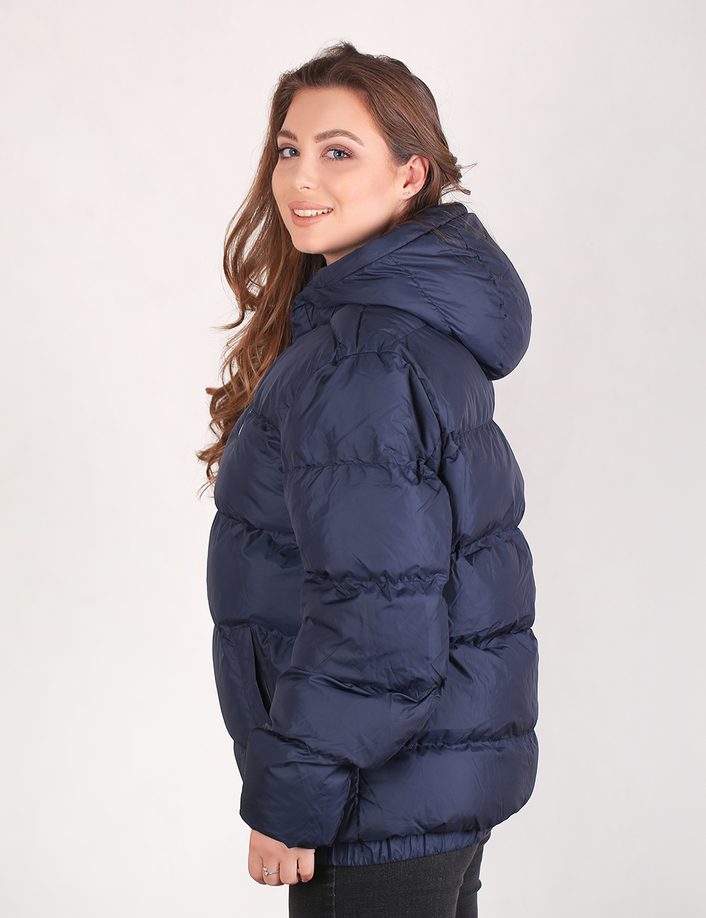 A woman wearing a navy blue hooded puffer jacket, shown from a side-back angle against a plain light gray studio background.