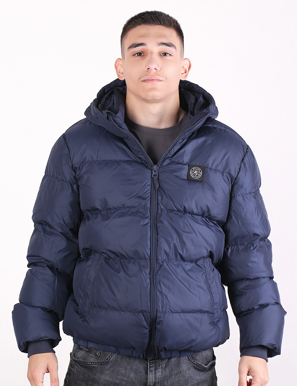 A man wearing a navy blue hooded puffer jacket with a zippered front and a circular chest patch, photographed against a plain white background.