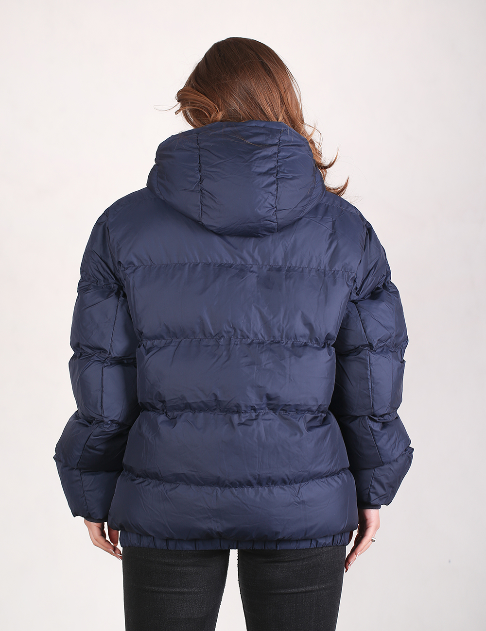 A woman viewed from the back wearing a navy blue puffer jacket with a hood and horizontal quilted stitching, paired with black pants, against a plain light background.