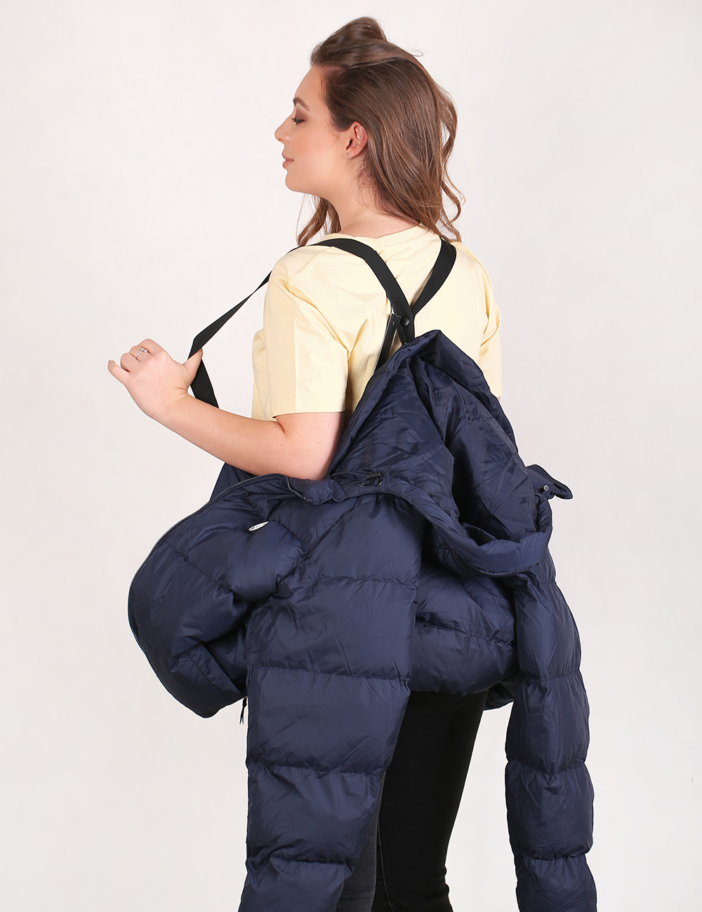 A woman shown from the back wearing a pale yellow t-shirt and black pants, carrying a navy blue puffer jacket over her shoulder against a plain white studio background.