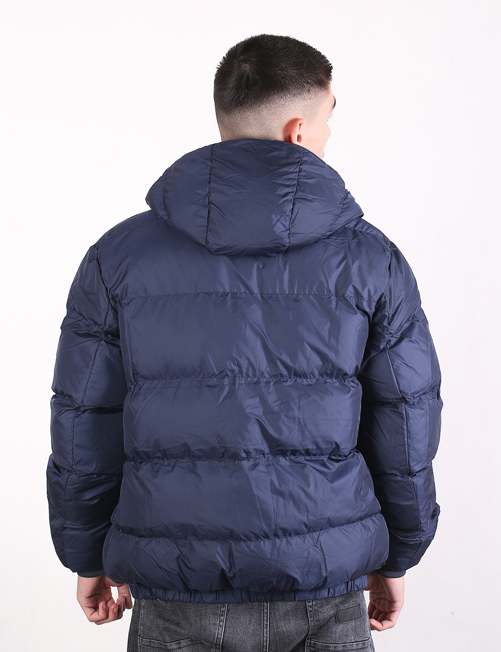 A man viewed from the back wearing a navy blue puffer jacket with horizontal quilting and a hood, paired with gray jeans, against a plain white background.