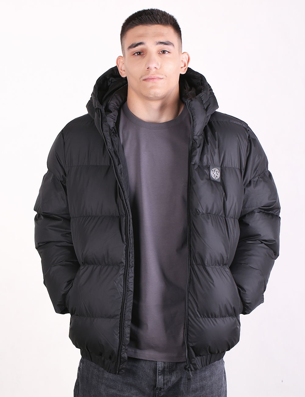 A man wearing a black hooded puffer jacket over a dark gray t-shirt and jeans, standing against a plain white background.