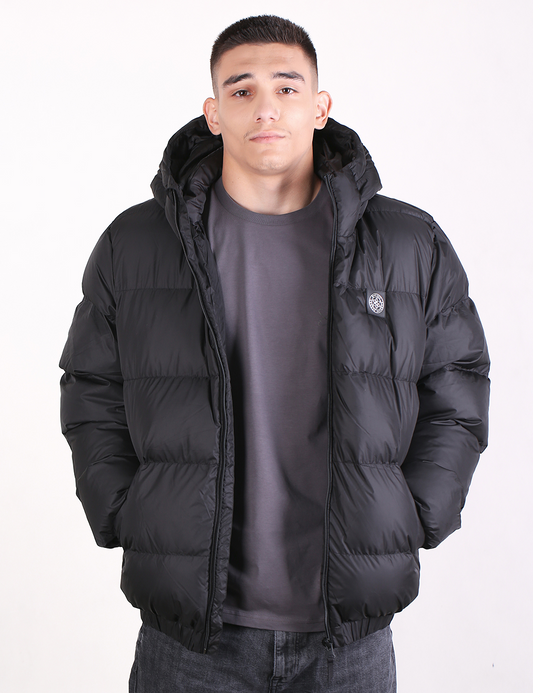 A man wearing a black hooded puffer jacket over a dark gray t-shirt and jeans, standing against a plain white background.
