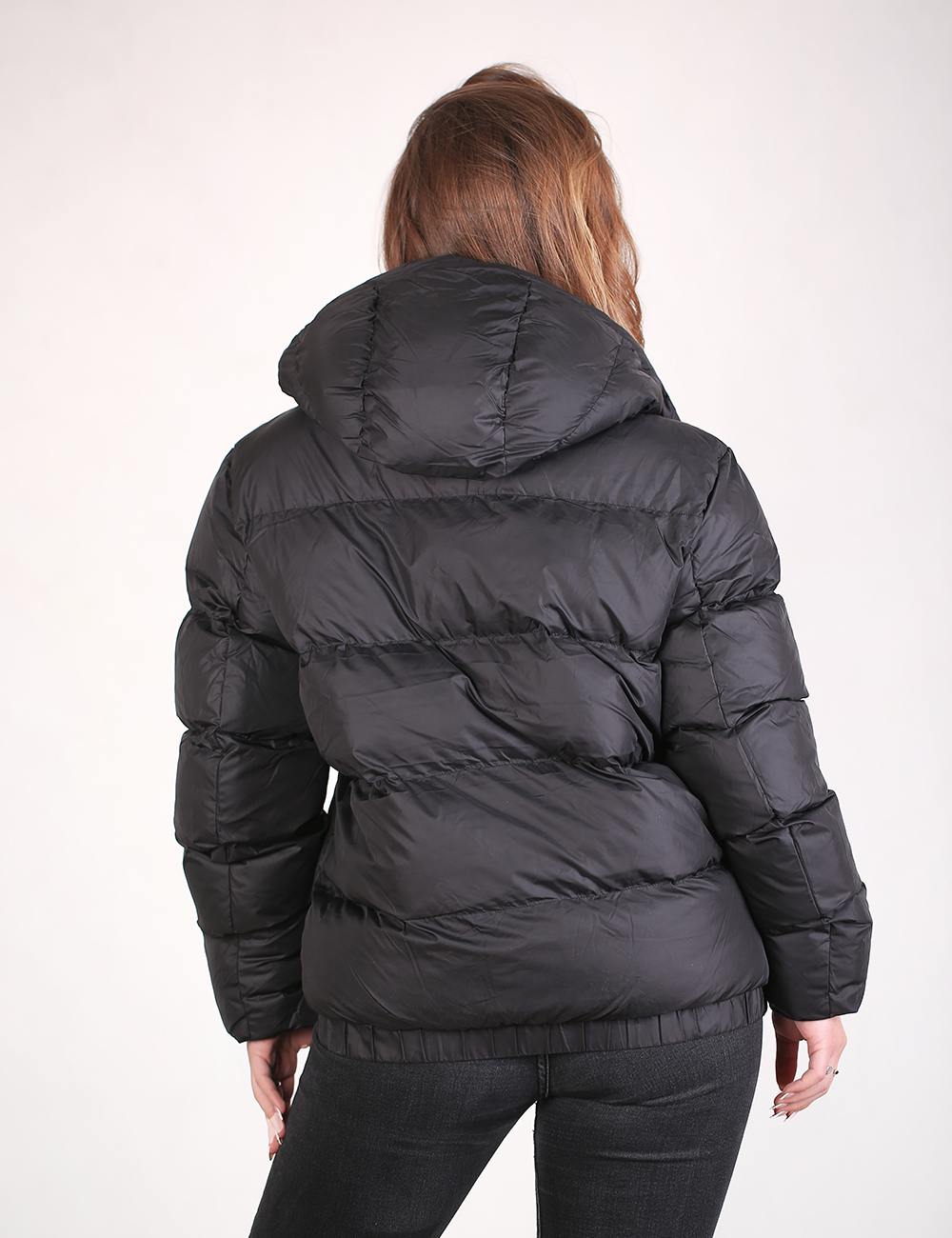 A woman viewed from the back wearing a black puffer jacket with horizontal quilting and a hood, paired with dark jeans, against a plain light background.