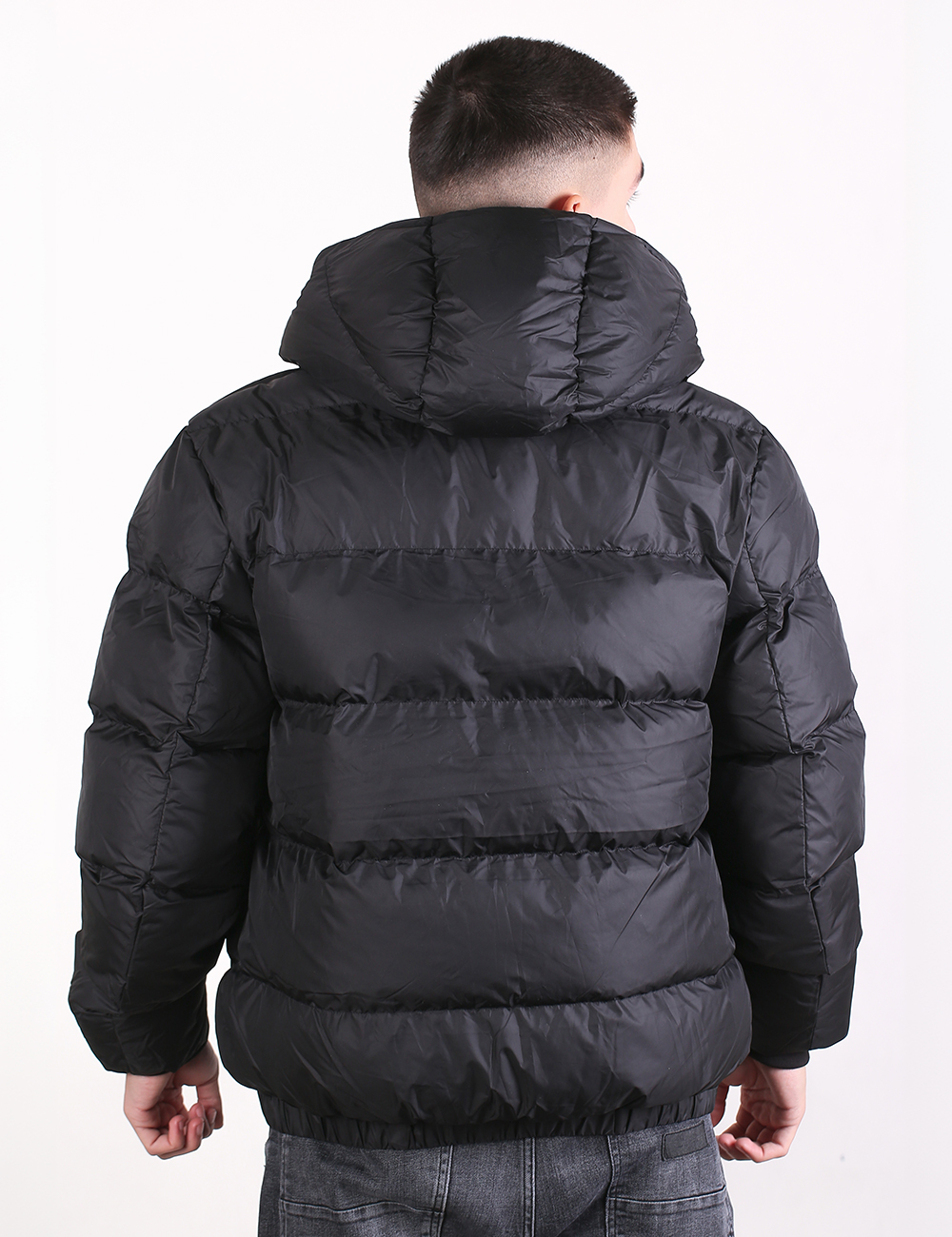 A man viewed from the back wearing a black puffer jacket with horizontal quilting and a hood, paired with gray jeans, against a plain white background.