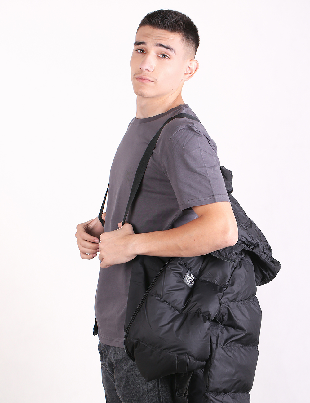 A man wearing a dark gray t-shirt and jeans, carrying a black puffer jacket over his shoulder with a black strap, set against a plain white studio background.