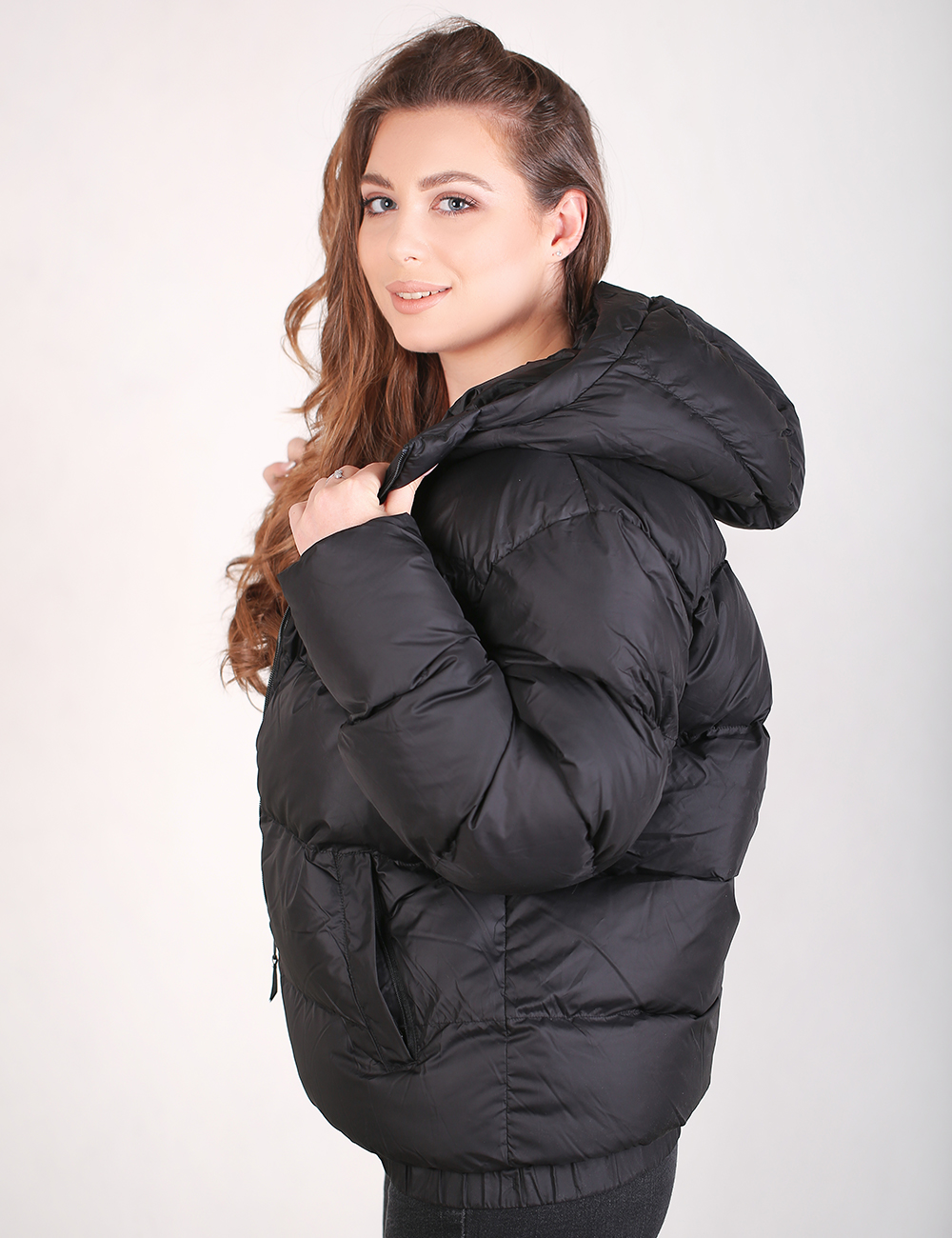 A woman wearing a black puffer jacket with a hood, posing against a light gray studio background.