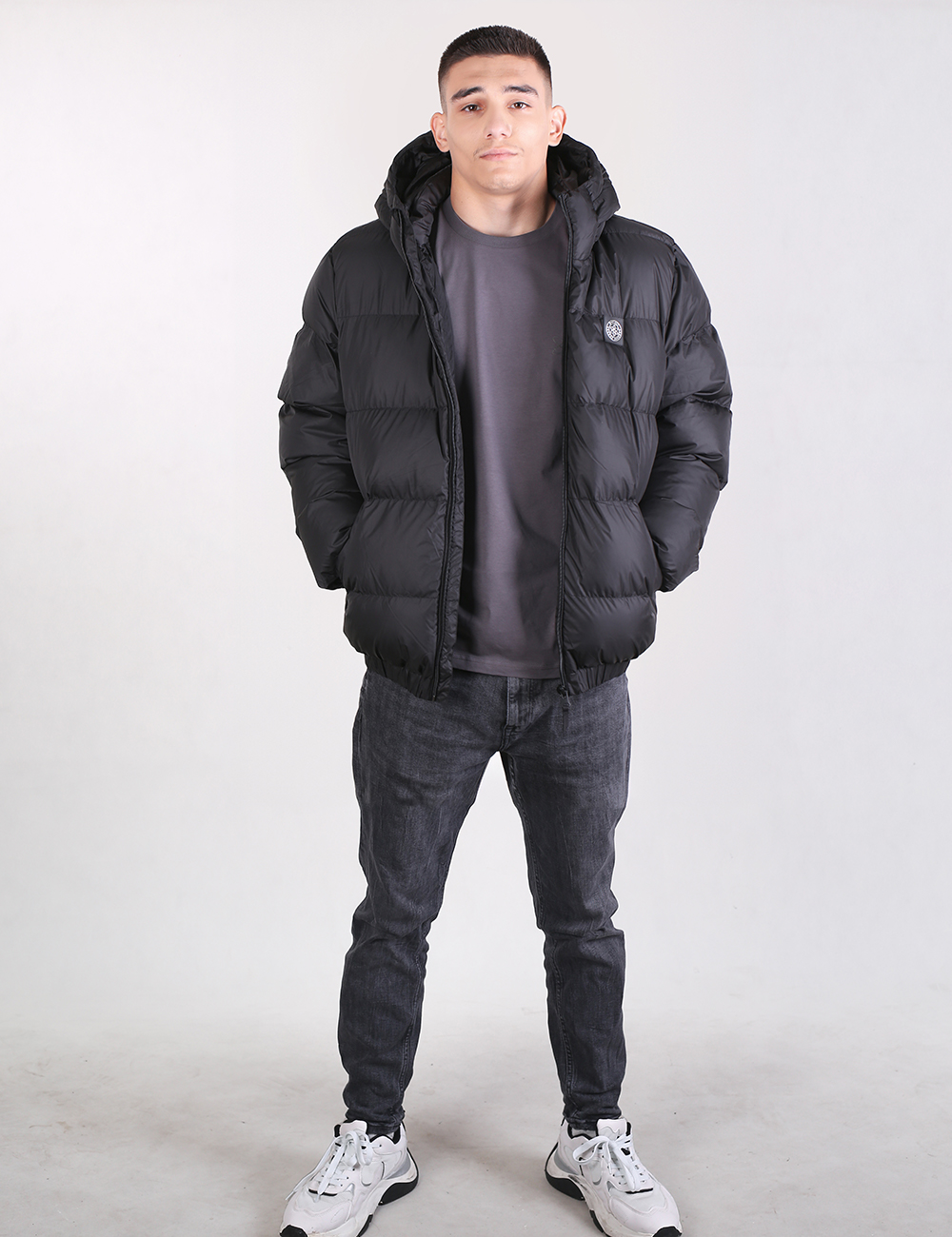 A man modeling a black hooded puffer jacket over a dark gray t-shirt, dark gray jeans, and white sneakers, standing against a light gray studio background.