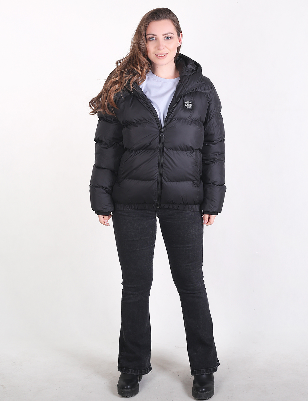 A woman modeling a black hooded puffer jacket with a front zipper, worn over a light gray top and black flared jeans, against a plain white studio background.