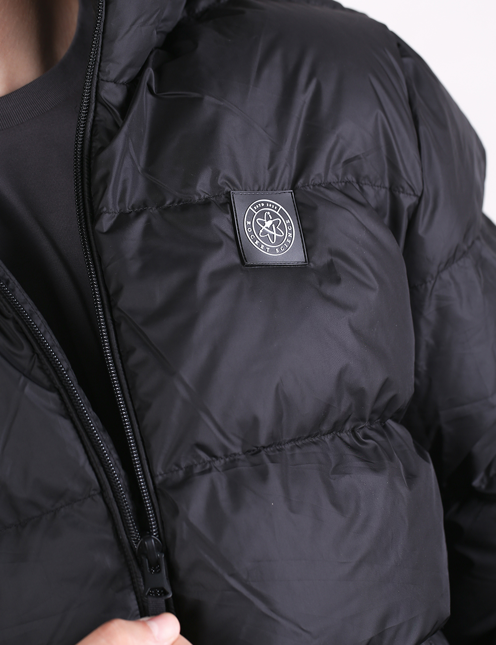 Close-up of a black puffer jacket with horizontal quilting, a black zipper, and a square black brand patch on the chest. A hand is visible at the bottom edge.