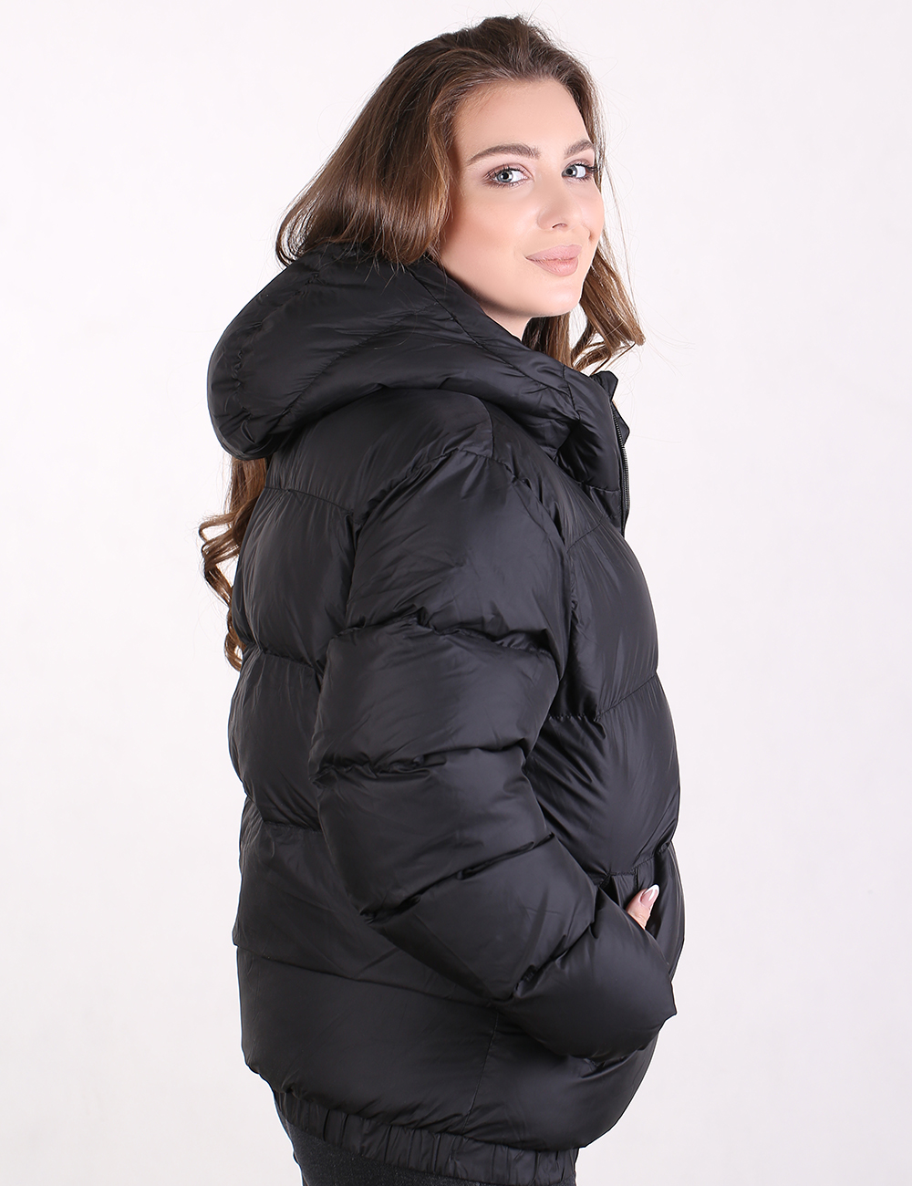 A woman wearing a black puffer jacket with a hood, shown from a side-back angle against a plain white studio background.
