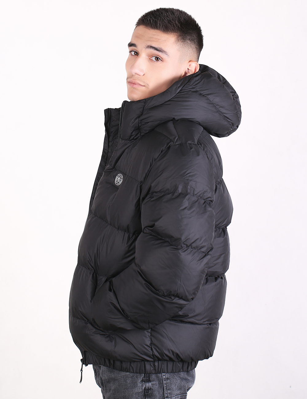 A man wearing a black hooded puffer jacket, shown in profile against a plain white studio background.