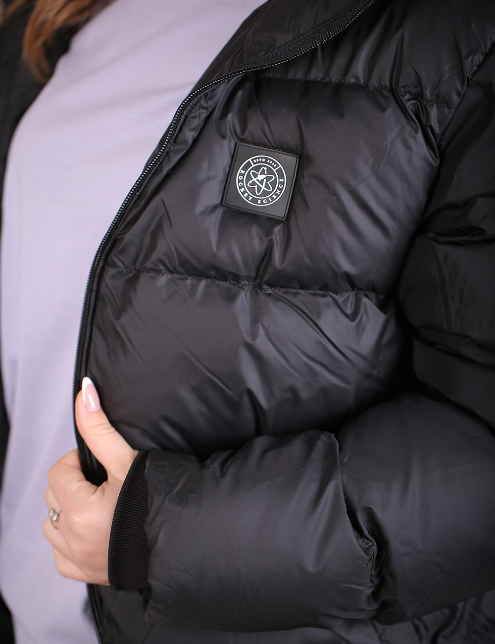 Close-up of a person wearing a black puffer jacket over a light lavender top, showing the jacket's quilted texture, zipper, and a circular brand patch on the chest.