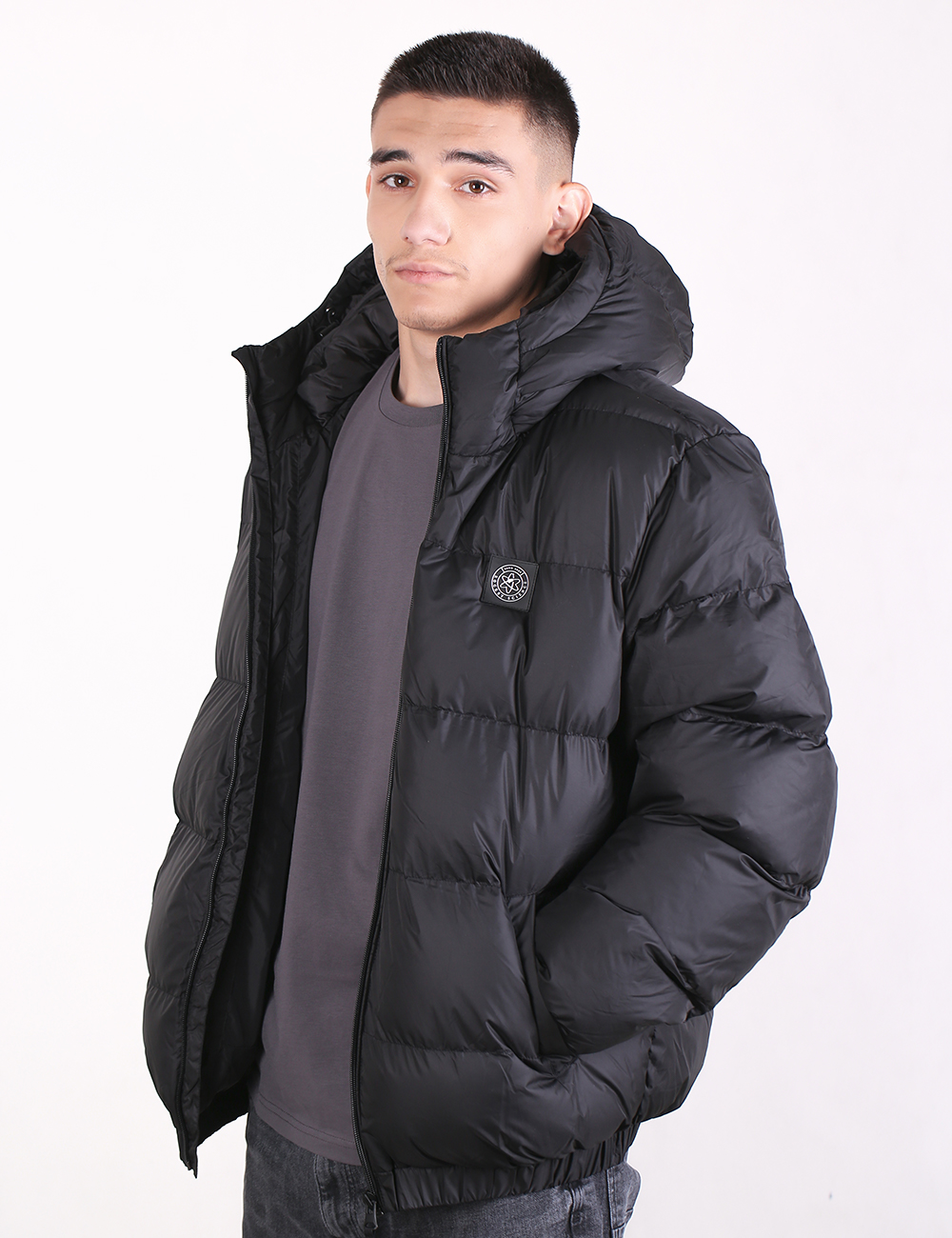 A man wearing a black hooded puffer jacket over a dark gray t-shirt, photographed against a plain white studio background.
