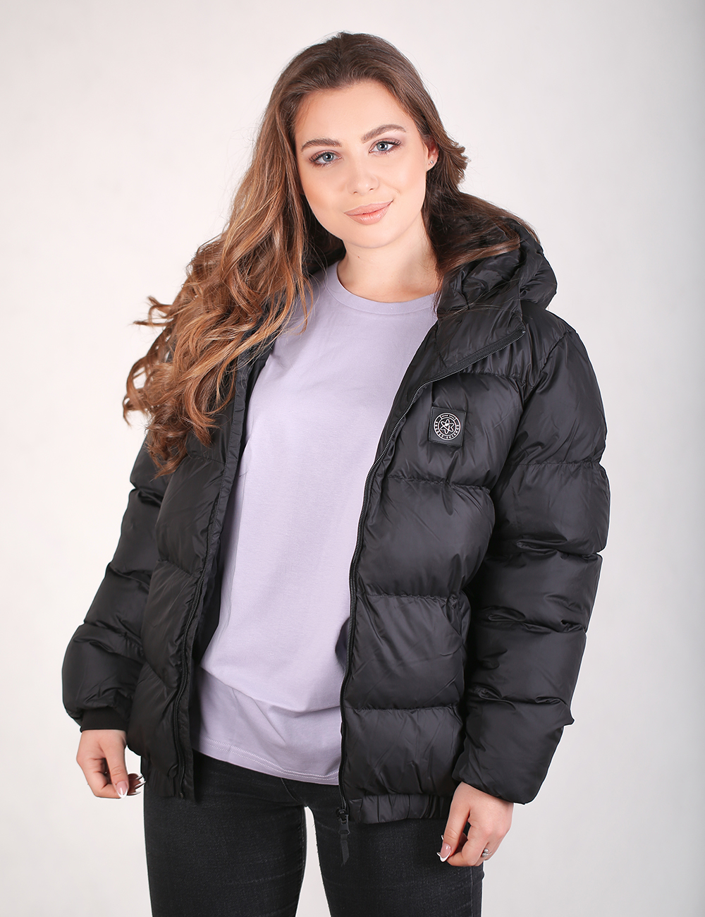 A woman wearing a black hooded puffer jacket over a light purple sweatshirt and black pants, standing against a plain light gray studio background.