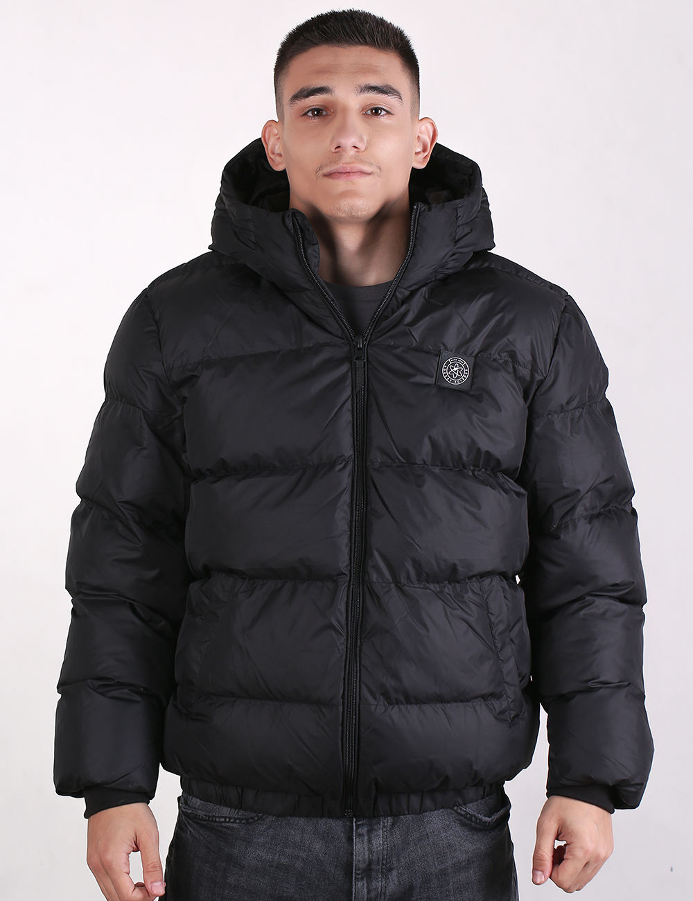 A man wearing a black hooded puffer jacket with horizontal quilting, a front zipper, and a circular chest logo, photographed against a plain light gray studio background.