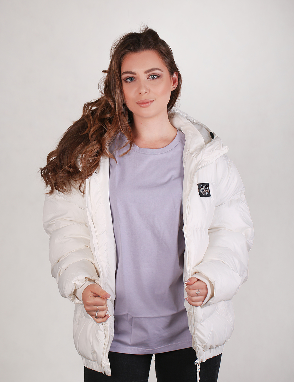 A young woman with long, wavy brown hair parted to the side looks directly at the camera with a subtle smile. She is wearing a bulky, white oversized puffer jacket with a black rectangular logo patch on the left chest, worn open over a light purple t-shirt and black pants. She holds the edges of the jacket with both hands, showing rings on her fingers, against a plain white background.