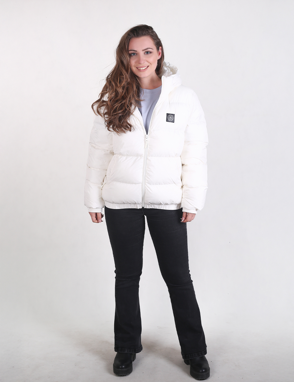 A woman modeling a white puffer jacket with a small square logo patch on the left chest, paired with black pants and black shoes, against a plain light gray studio background.