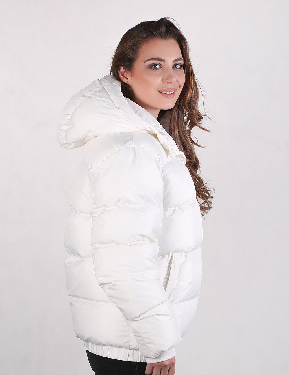 A woman wearing a white puffer jacket with a large hood, shown from a side-back angle against a plain light gray studio background.