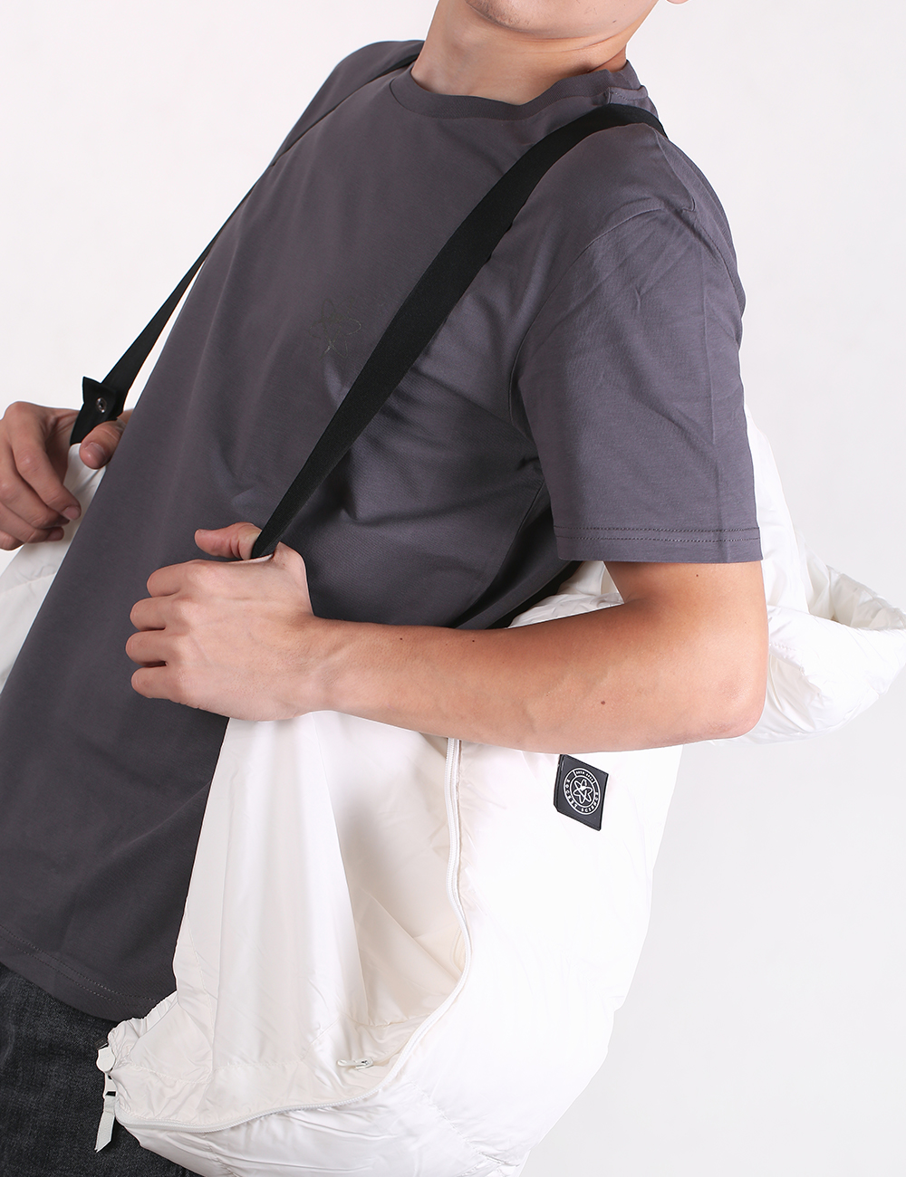 Person wearing a gray shirt with a black strap over their shoulder, holding a white bag.