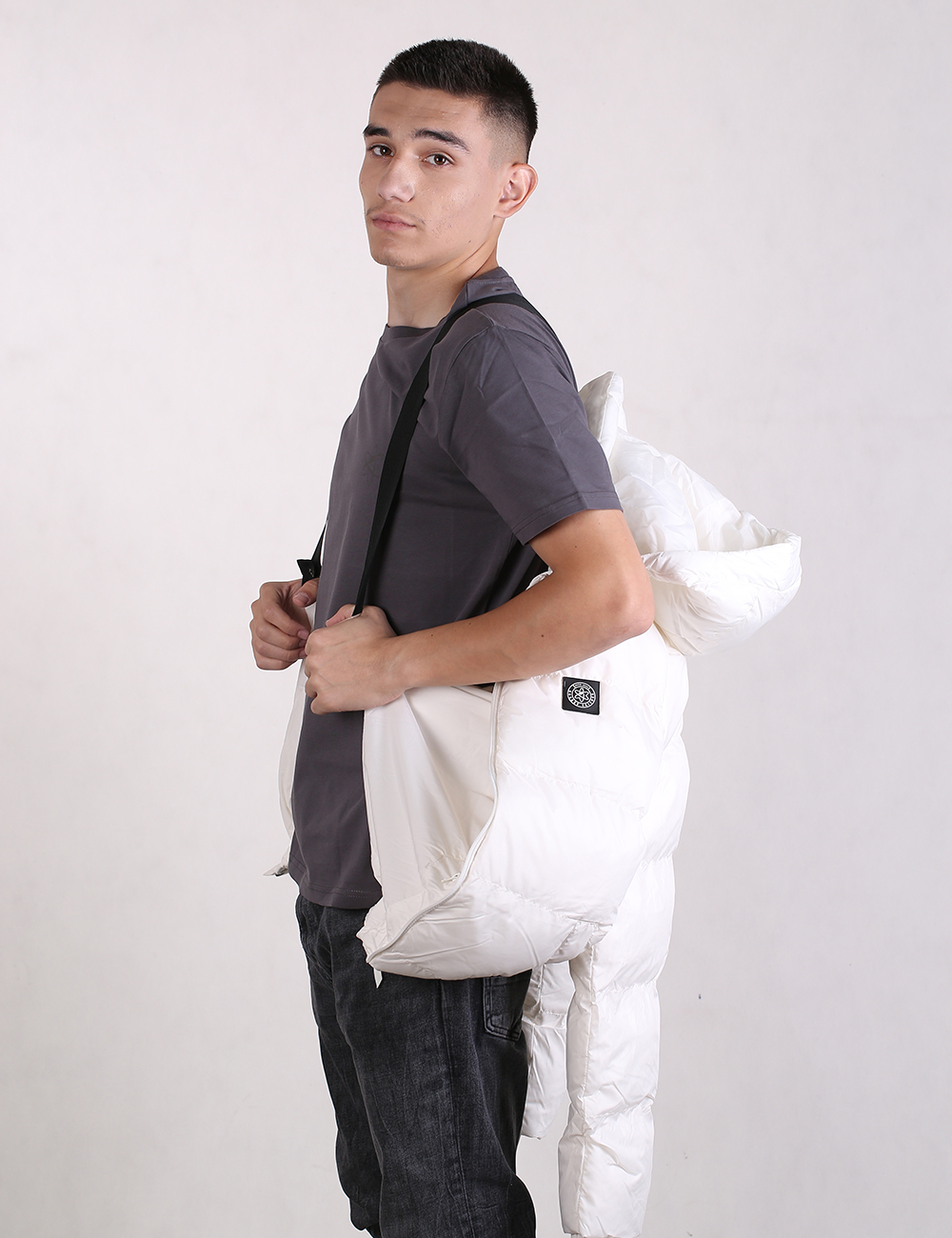 A man wearing a dark gray t-shirt and dark jeans is shown from the side, carrying a white puffer jacket over his shoulder on a plain light gray studio background.