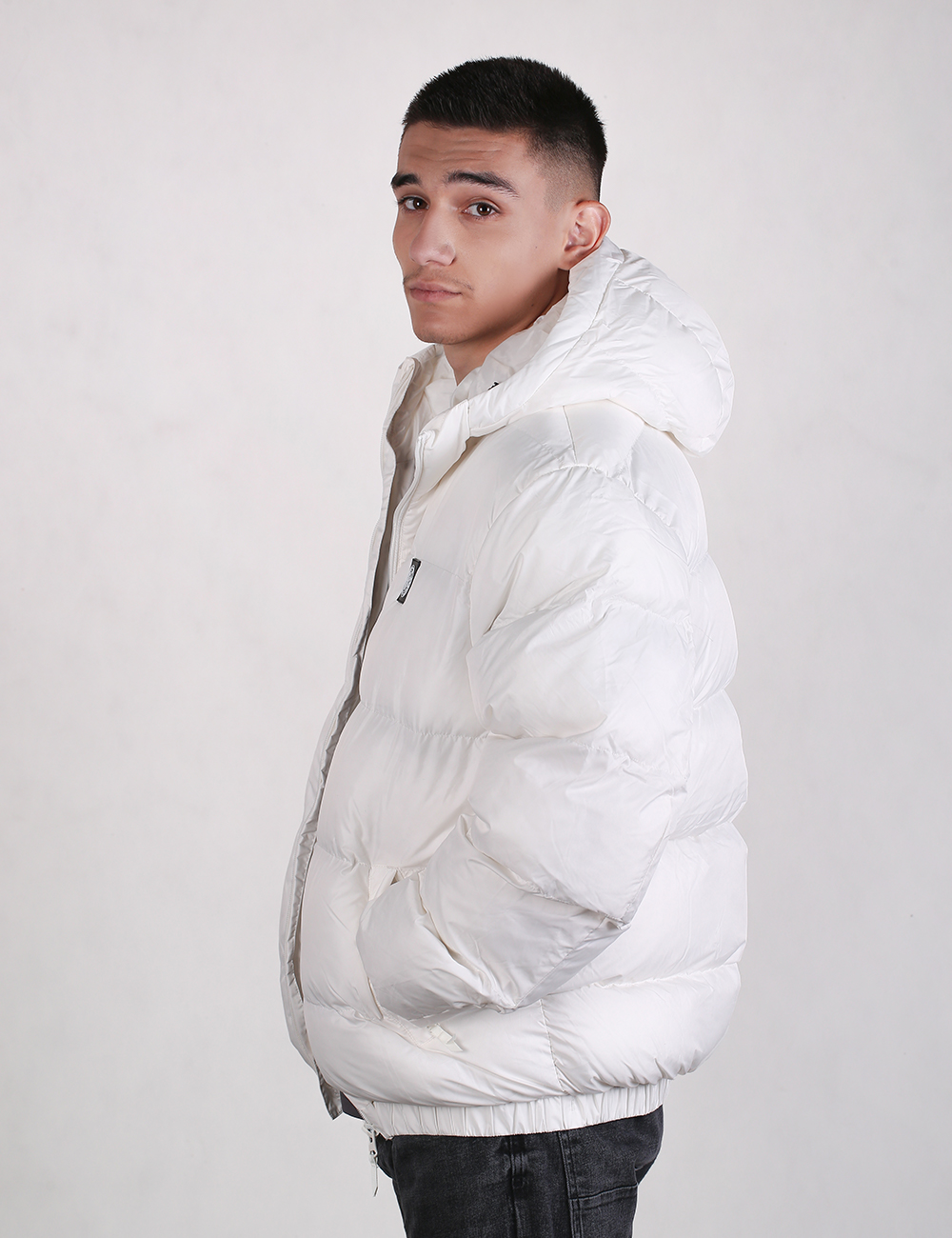A man stands in side-profile, looking back toward the camera over his left shoulder. He has short, dark hair and dark eyes. He is wearing a thick, oversized, white puffer jacket with a hood and black jeans. The background is a solid, light grey.