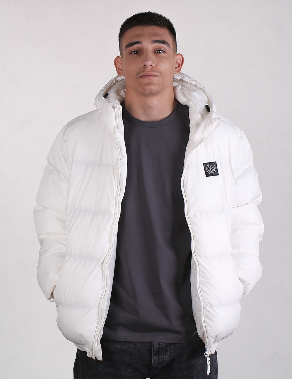 A man wearing a white puffer jacket with a black chest logo patch over a dark gray t-shirt and black jeans, standing against a light gray studio background.