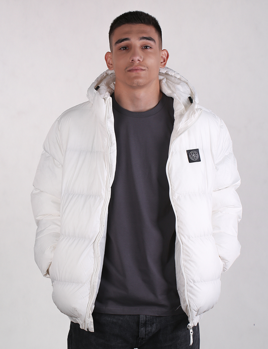 A man wearing a white puffer jacket with a black chest logo patch over a dark gray t-shirt and black jeans, standing against a light gray studio background.