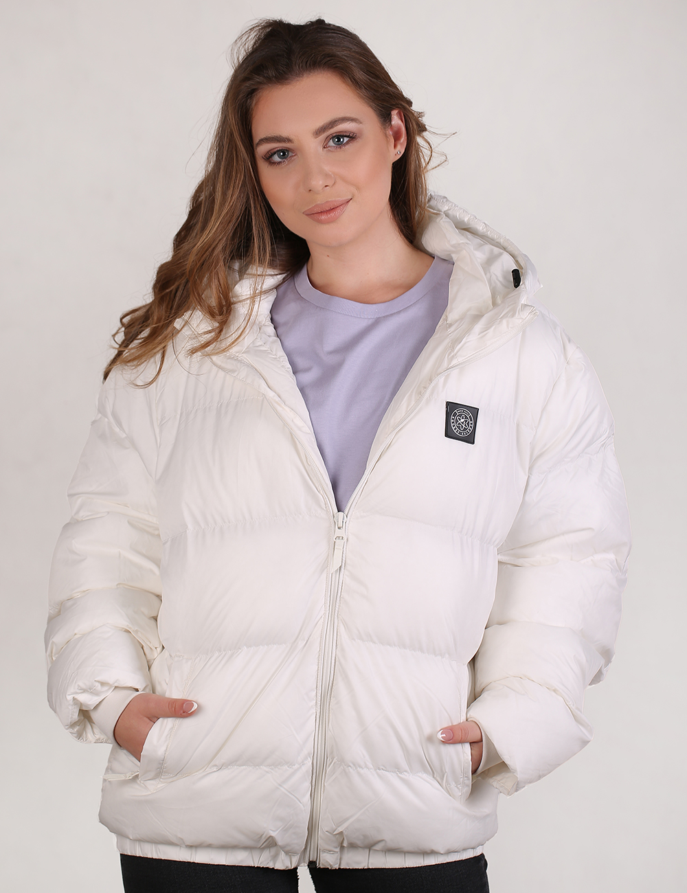 A woman wearing a white puffer jacket with a zip-up front, quilted padding, and a small black brand patch on the chest, over a light purple t-shirt.
