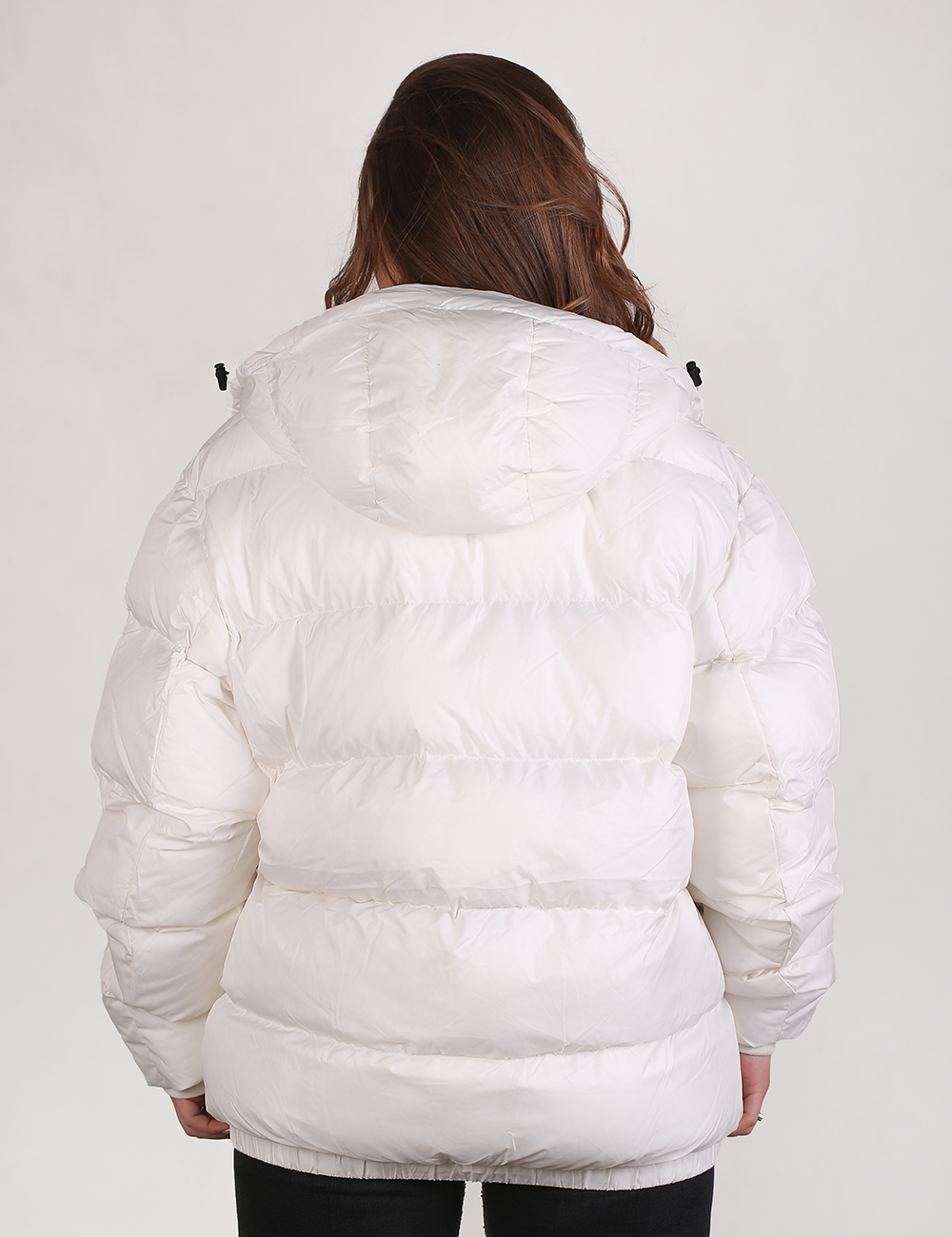 Rear view of a person with long brown hair wearing a thick, oversized white puffer jacket with a hood. The jacket features horizontal quilted stitching and black toggles on the hood drawstrings. The person is wearing black pants and standing against a plain light gray background.