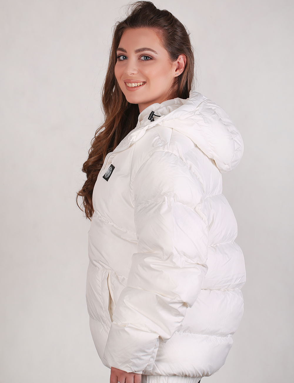 A woman wearing a white puffer jacket with a hood, shown from a side-back angle against a light gray studio background.