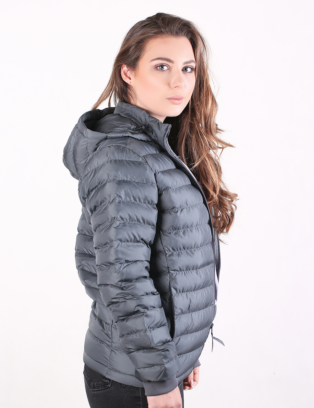 A woman wearing a dark gray hooded puffer jacket, shown from a side-back angle against a plain white studio background.