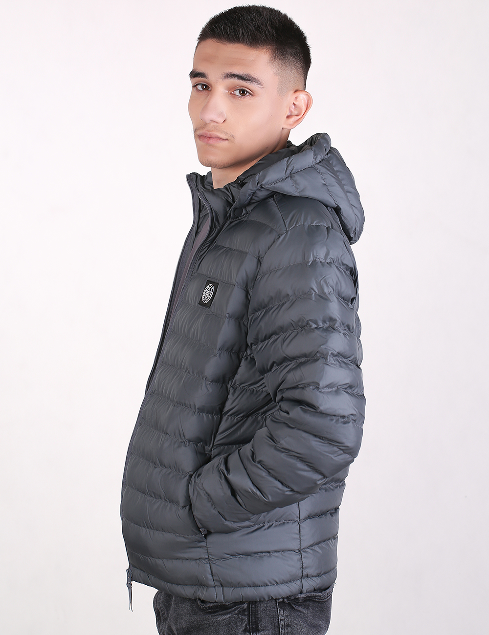 A man wearing a dark gray hooded puffer jacket, shown in profile against a plain white studio background.