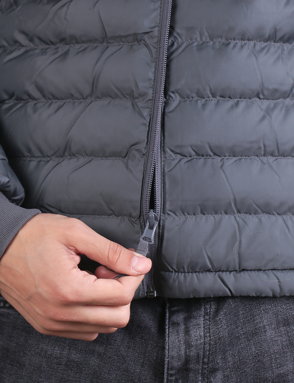 A close-up shot focuses on a person's hand pulling the zipper pull tab of a dark grey, channel-quilted puffer jacket. The individual is wearing grey jeans, and the zipper is partially undone near the bottom hem of the coat.