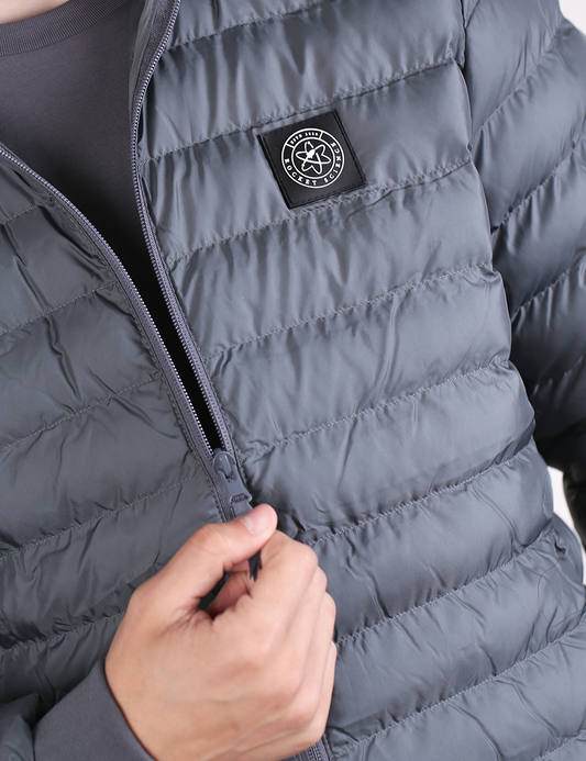 A close-up photograph of a person wearing a grey puffer jacket with a front zipper partially open. On the left chest area, there is a square, black stitched patch featuring a white circular logo that includes an atom symbol with the text "Rocket Science." 