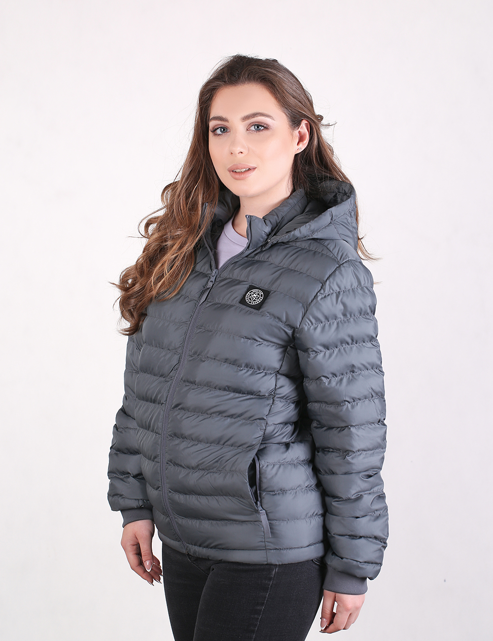 A young woman with long, wavy brown hair poses from the waist up, looking directly at the camera with a subtle smile. She is wearing a grey, hooded puffer jacket with thin horizontal quilting and a small, round black logo patch on the left side of the chest. She is also wearing dark trousers, and the background is a solid, clean white.