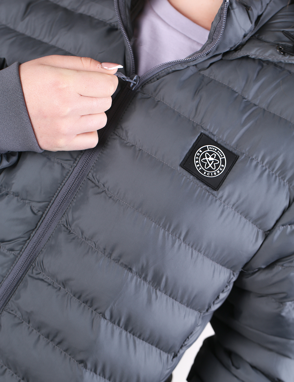 Close-up of a person wearing a dark gray puffer jacket, pulling the zipper with one hand. A square black brand patch on the chest reads 'Rocket Science'.